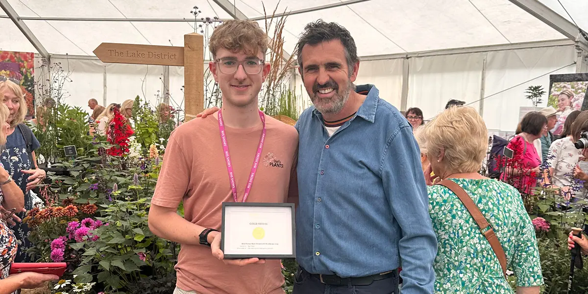 Two men stand together inside a large tent full of people and flowers. One young man holds a certificate and wears a lanyard, while the older man smiles with an arm around him. A Lake District sign is visible behind them.