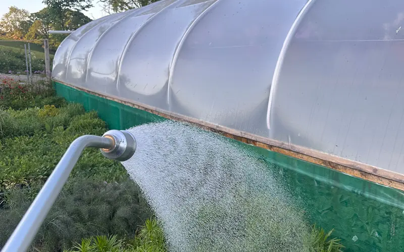 A watering can spout sprays water over green plants next to a large greenhouse with a translucent plastic cover.